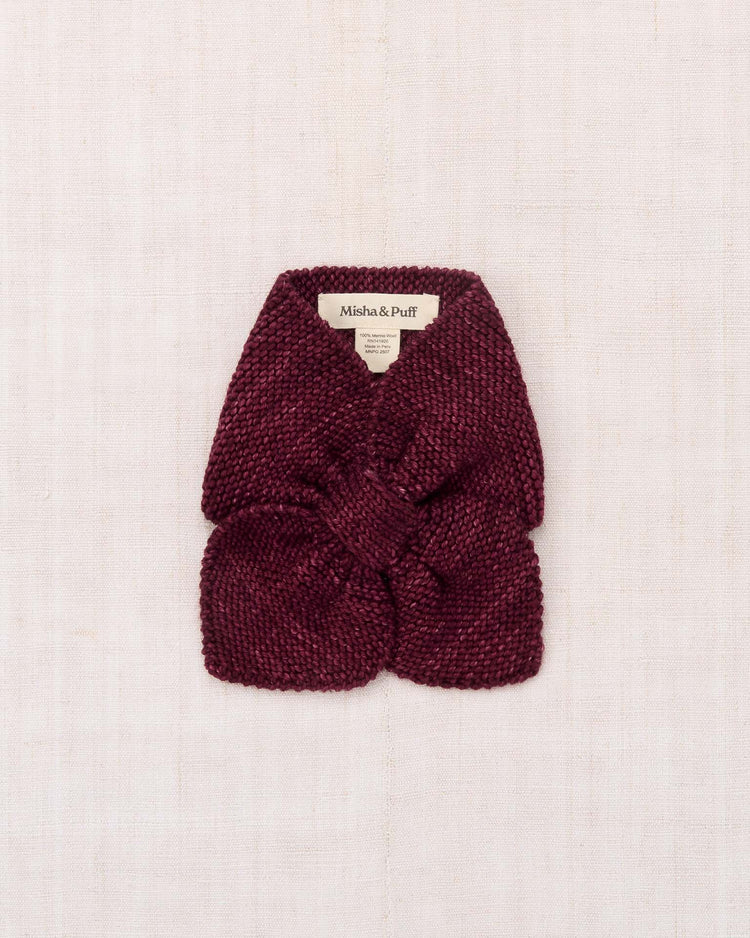 A deep burgundy sledding scarf in cambridge, hand-knit from soft Merino wool with a charming bow detail, is neatly folded on a light beige textured background. A misha + puff label is visible at the top.