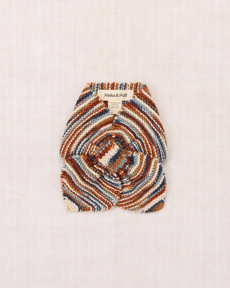 The “sledding scarf in neriage space dye” by misha + puff is a multicolored, knitted neck warmer made from hand-dyed Merino wool with diagonal stripes in brown, cream, blue, and orange. It is folded neatly with a misha + puff label at the top and placed on a light beige fabric background.