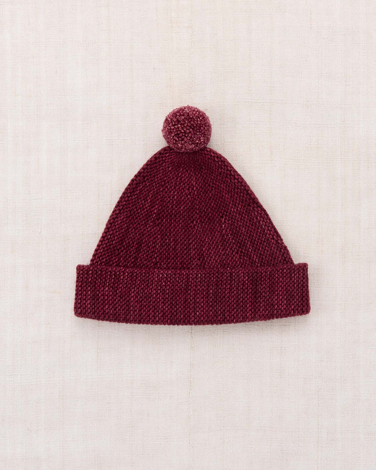The garter hat in cambridge by misha + puff is a maroon knitted beanie, crafted from soft Merino wool with a folded brim and a playful pom-pom on top. Laid flat on a light beige fabric background, the hat highlights its cozy design and luxurious texture.