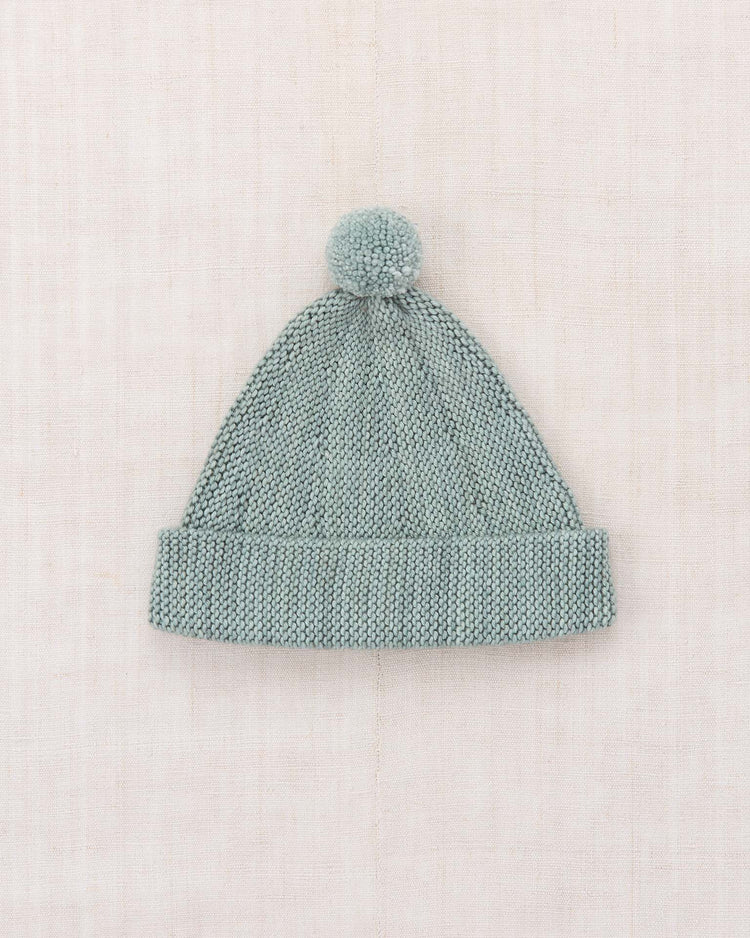 A light gray garter hat in onsen by misha + puff, crafted from Merino wool and featuring a fold-over cuff and a playful pom-pom on top, is laid flat on a light neutral fabric background. The adjustable brim ensures both comfort and style for any occasion.