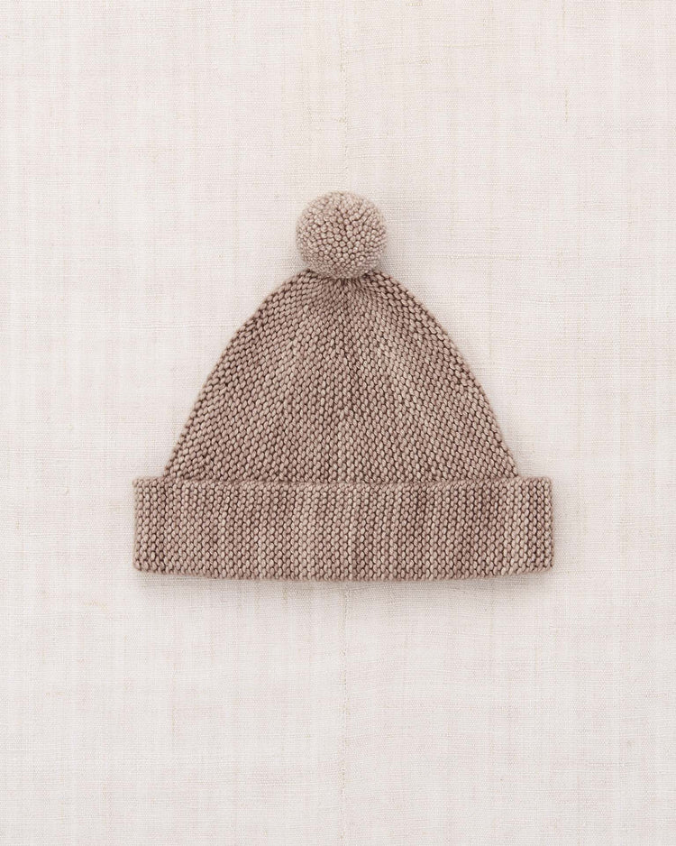A garter hat in turtledove by misha + puff, crafted from knitted Merino wool with a folded, adjustable brim and topped with a pompom, displayed flat on a light, textured background.