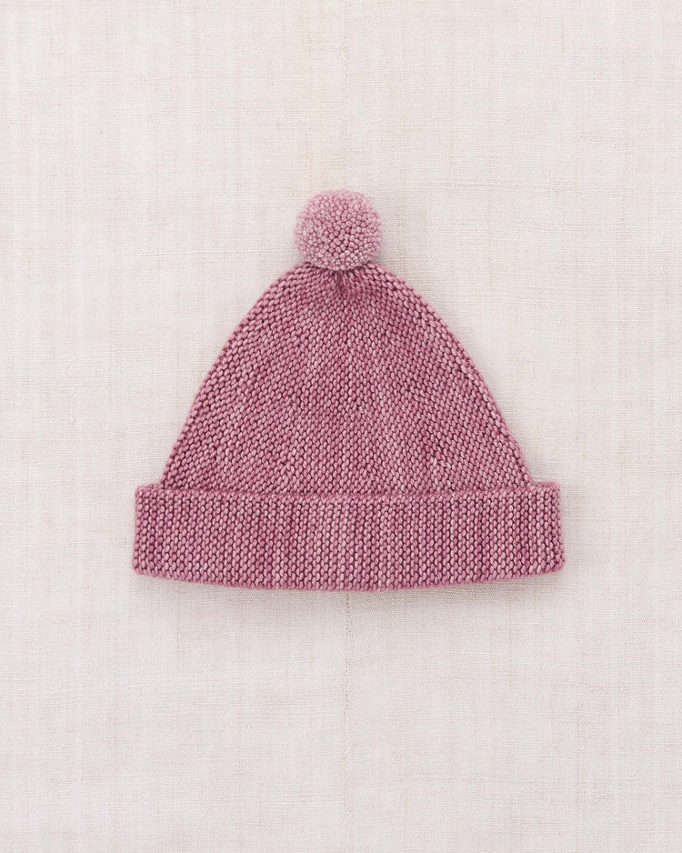 The “garter hat in antique rose” by misha + puff, a knitted Merino wool hat featuring a folded, adjustable brim and a playful pom-pom on top, is laid flat on a light textured background.