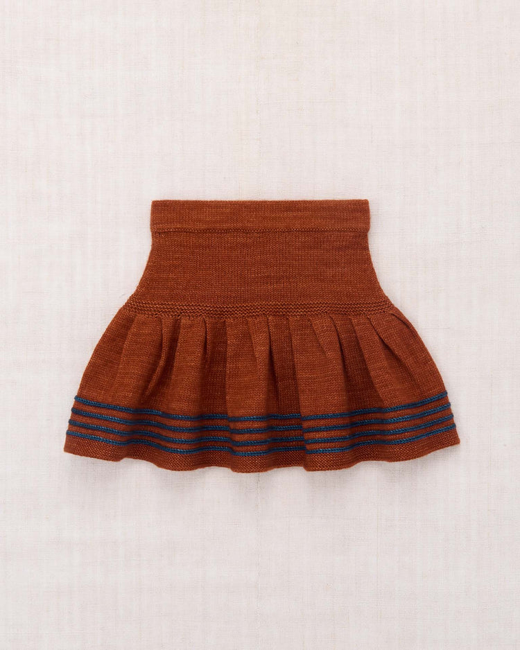 The judy skirt in sabi by misha + puff, a brown knitted skirt accented with blue horizontal stripes near the hem and subtle hand-worked details, is shown laid flat on a light textured background.