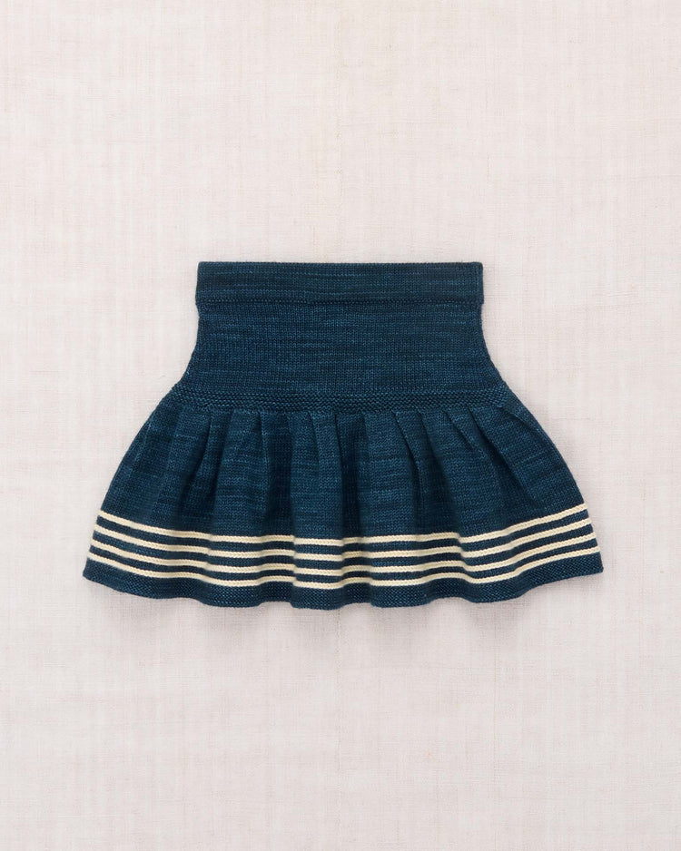 The judy skirt in marine blue by misha + puff features a high waistband, pleated design, and three cream-colored horizontal stripes near the hem. Crafted from dark blue Merino wool and displayed on a light fabric background, this skirt showcases a flowing skating silhouette with hand-worked details that highlight both movement and exceptional craftsmanship.