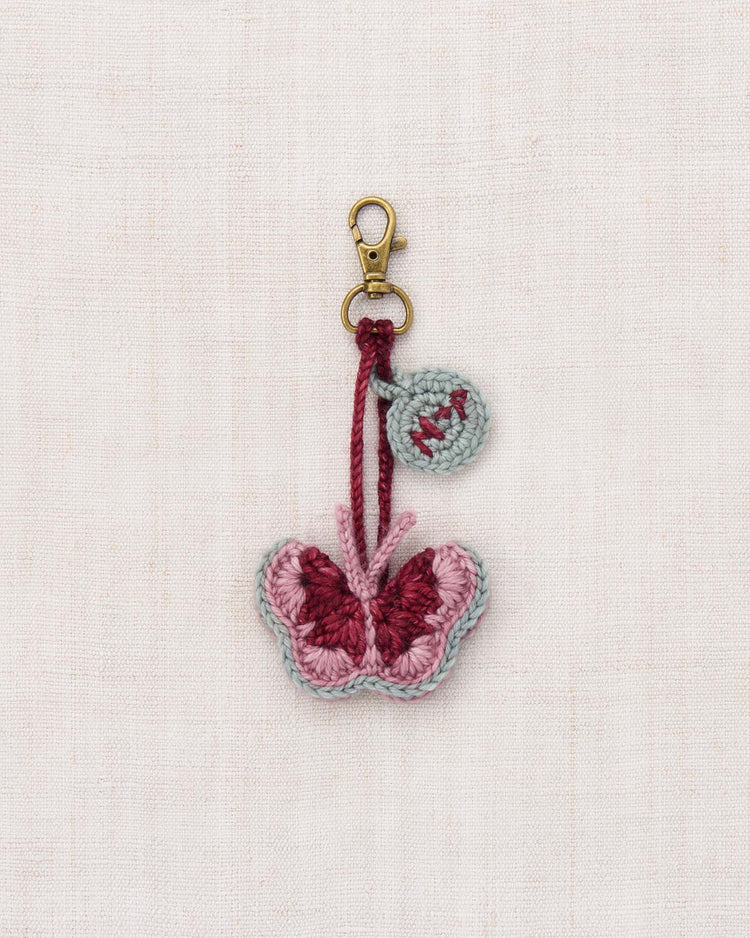 A mariposa bag charm in antique rose by misha + puff—a hand-crocheted butterfly keychain in pink and burgundy, featuring a round tag and brass clasp, displayed on a light fabric background—perfect as a merino wool bag charm or unique accessory for your favorite bag.