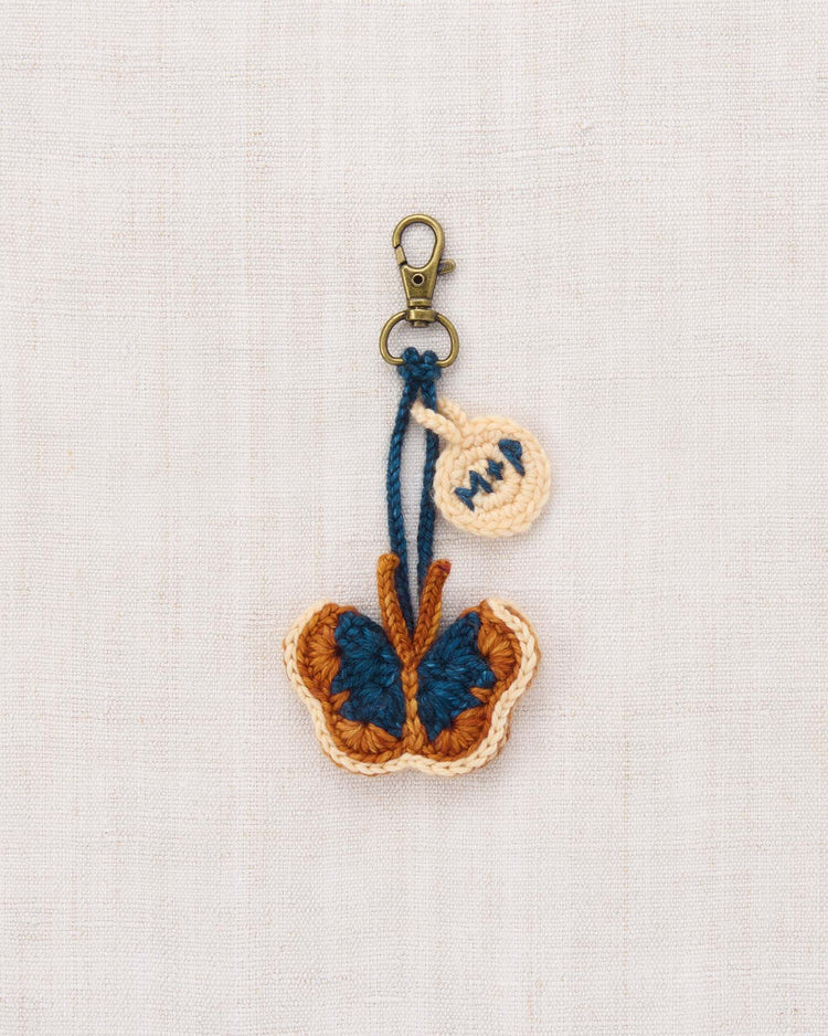 The mariposa bag charm in fox by misha + puff is a hand-crocheted butterfly keychain crafted from blue, brown, and beige merino wool yarn. Featuring a metal clasp and a small beige tag, it makes a charming accessory for your bag. Shown here on a light fabric background.