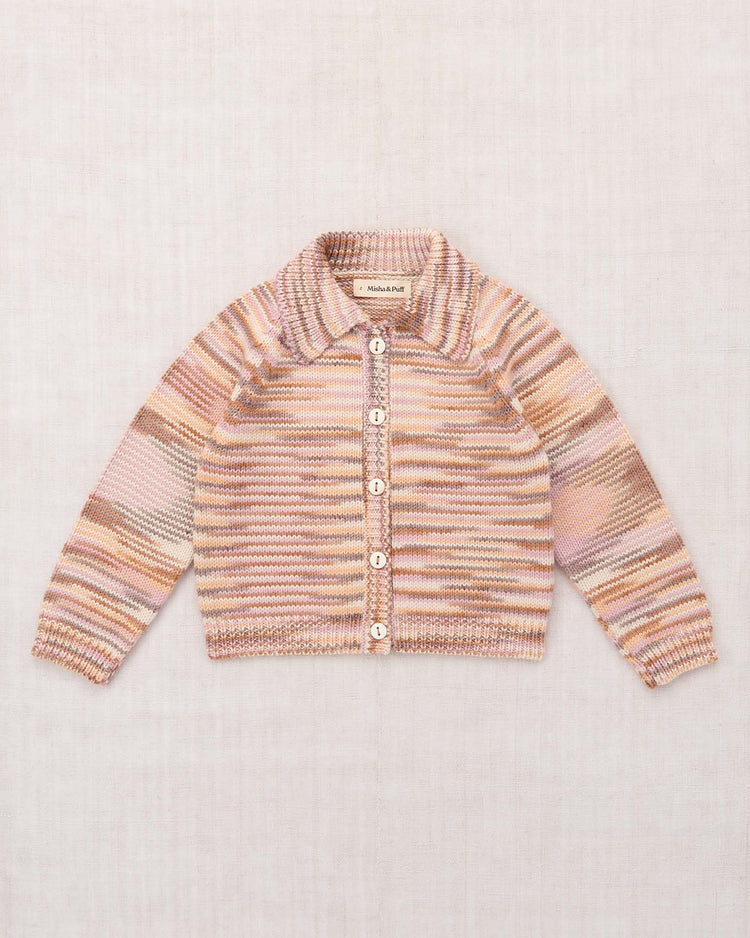 The motley cardigan in mochi space dye by misha + puff, a pastel-hued, striped knit crafted from soft Merino wool with long sleeves and six buttons, is laid flat on a light textured background.