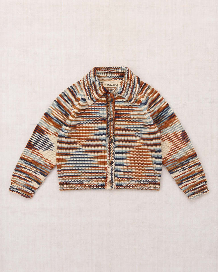 Introducing the motley cardigan in neriage space dye by misha + puff—a vintage-inspired cardigan with a zip front, showcasing a geometric striped pattern in brown, beige, orange, and blue hues. Expertly crafted from soft Merino wool and presented against a light fabric background.