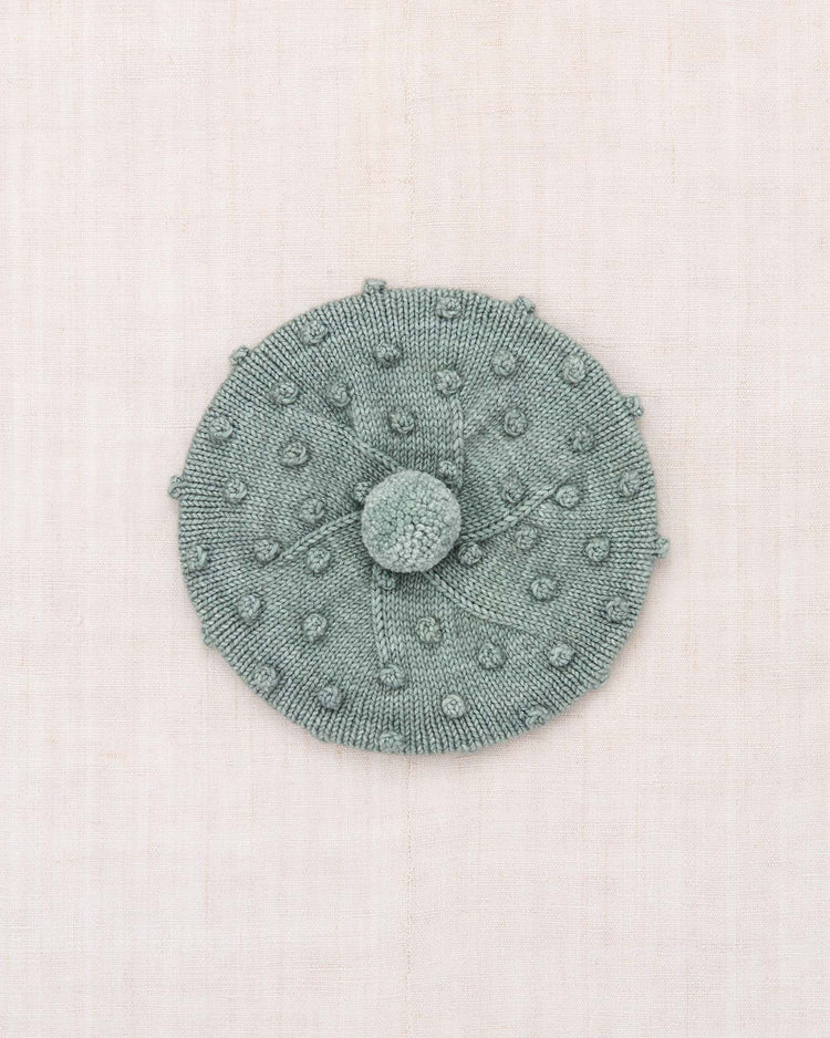 The popcorn beret in onsen by misha + puff is a light green hand-knit Merino wool beret featuring a textured popcorn stitch polka dot pattern and a playful pom-pom on top, shown flat against a light beige fabric background.