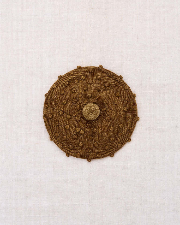 The misha + puff popcorn beret in nutmeg, featuring a round, hand-knit brown design with raised bumps from the popcorn stitch and a central stem, is centered against a plain off-white background.