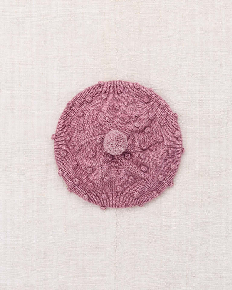 The misha + puff popcorn beret in antique rose—a hand-knit hat featuring a central pom-pom and a textured bobble pattern—is laid flat on a light beige fabric background.