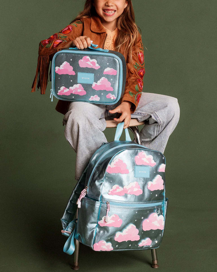 A child sits on a stool holding a blue lunchbox with pink clouds, which matches the State Bags Kane Backpack in Clouds, against a plain green background.