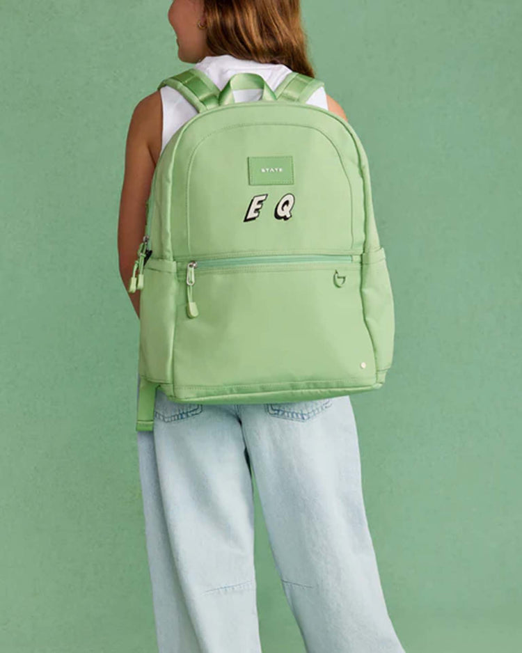 A person wearing a sleeveless white top and light blue jeans stands with their back to the camera, carrying a large kane double pocket backpack in green by state bags against a green background.