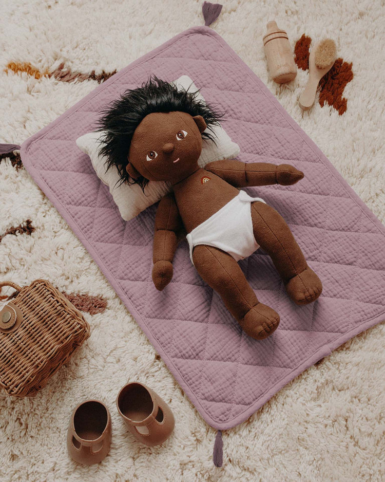 A dinkum dolls plush doll with dark brown skin and black hair rests on the strolley bedding set in lavender atop a cream rug, surrounded by brown shoes, a wicker basket, wooden bottle, and brush.