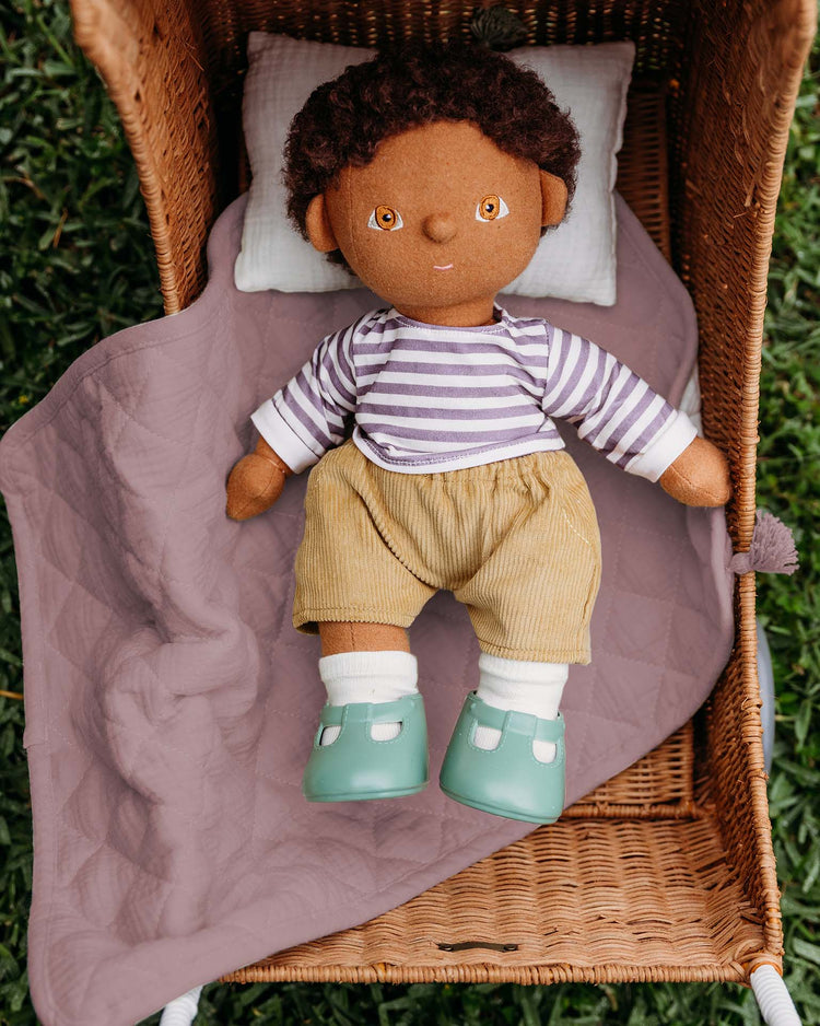 A dinkum dolls soft doll with brown skin and curly hair, wearing a striped shirt and tan shorts, lies on the strolley bedding set in lavender inside a wicker bassinet placed on green grass.