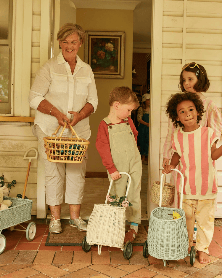 An older woman holding a dinkum dolls tarry basket in wheat stands at a doorway with three children, each with a kids storage basket or an Olli Ella wagon. They are smiling and enjoying a spring or Easter activity.