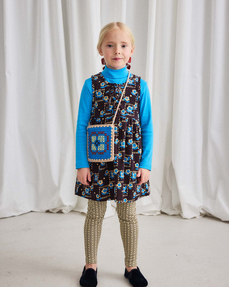 A young girl stands in front of white curtains, wearing the misha + puff turtleneck in blue smoke, a patterned dress, beige patterned leggings, black shoes, and a blue crocheted shoulder bag.