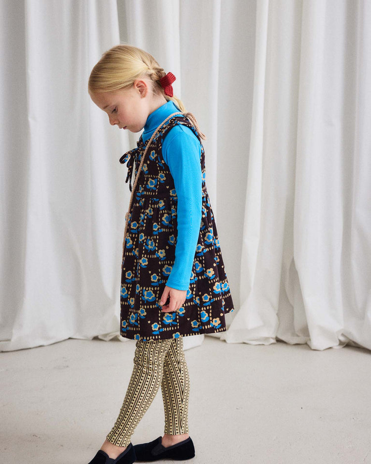 A young girl with blonde hair in braids stands indoors, looking down, with white curtains in the background. She is wearing the misha + puff turtleneck in blue smoke, a patterned dress, and geometric leggings.