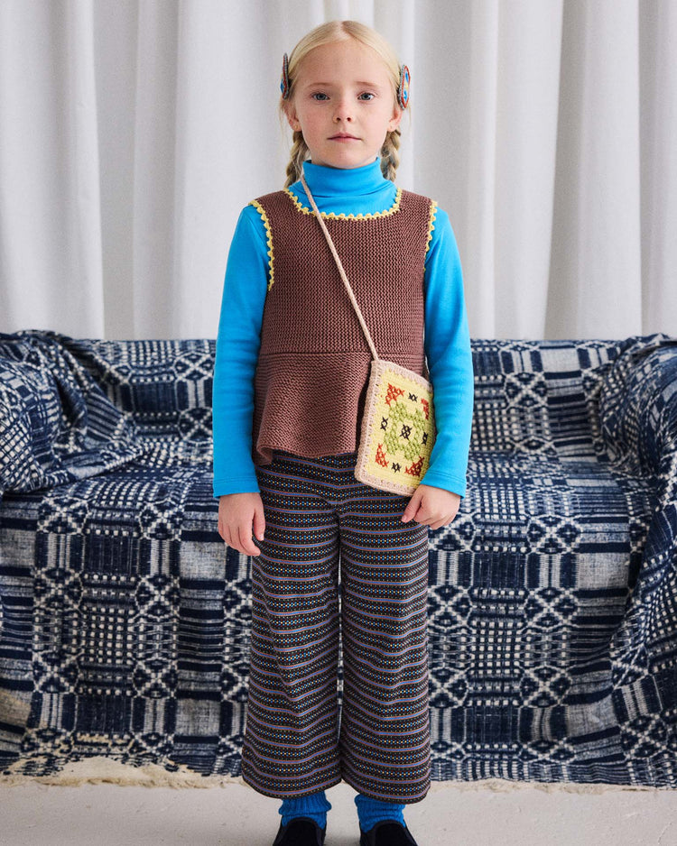 A young child with blonde hair stands indoors, wearing the misha + puff turtleneck in blue smoke made from organic Pima cotton, paired with a brown knitted vest, striped pants, and a small crocheted purse. A patterned blue and white couch and white curtains are visible in the background.