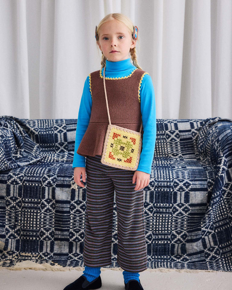 A young child stands indoors, wearing the misha + puff turtleneck in blue smoke layered beneath a brown knit vest, paired with striped pants and holding a small crocheted bag. A couch behind features a blue and white geometric patterned cover.