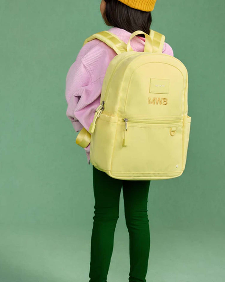 A child wearing a yellow beanie, pink sweater, green pants, and the Kane Mini Backpack in Butter Yellow from State Bags stands facing away against a green background.