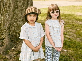 Two young girls standing outdoors near a tree, wearing coordinating light blue outfits with floral patterns.