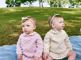 Two young children sitting on a blanket in a park wearing the new Misha + Puff Spring Collection sweaters and hair accessories.