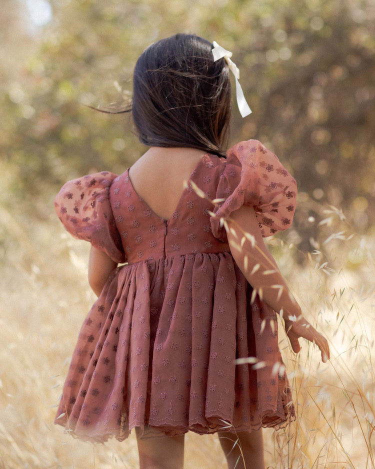 A young girl with long dark hair and a white bow, wearing the noralee sofia dress in spice floral with balloon puff sleeves, stands in a sunlit field of tall, dry grass, facing away from the camera.