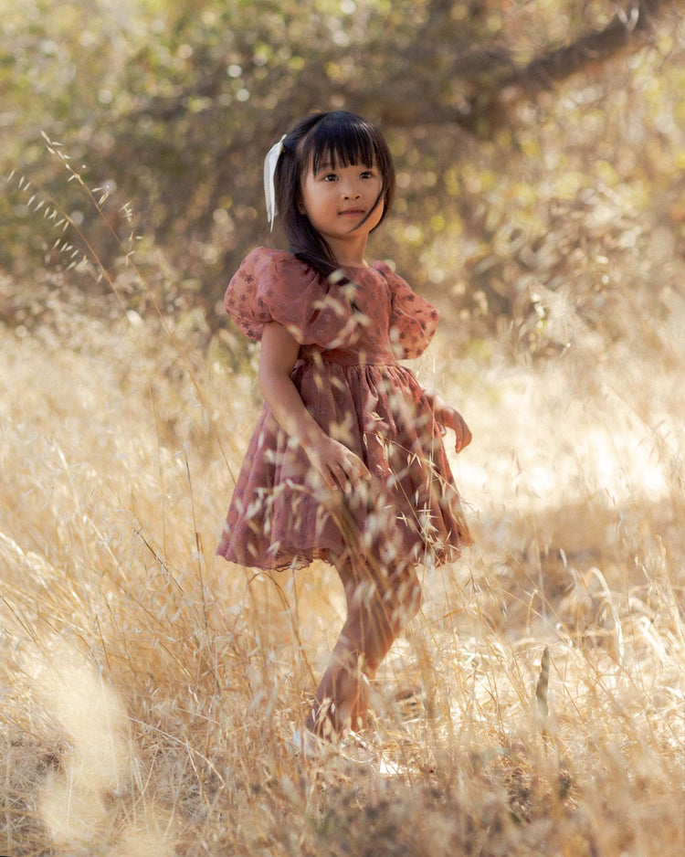 A young girl in the noralee sofia dress in spice floral, featuring balloon puff sleeves, stands barefoot in a sunlit grassy field surrounded by tall, dry grasses and trees. Her long dark hair is tied back with a white ribbon.