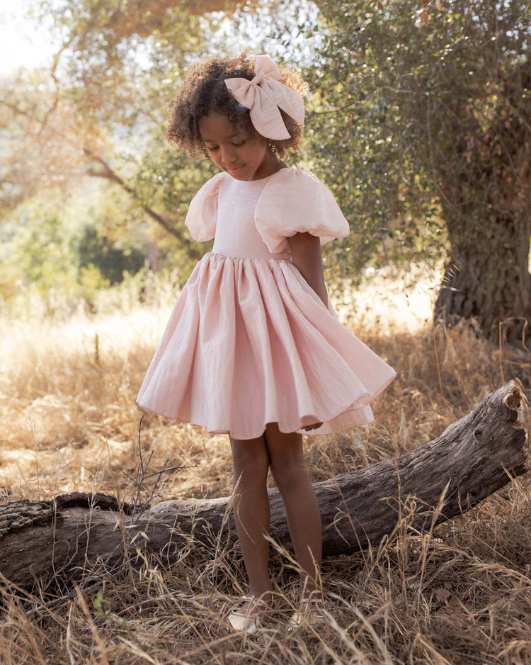 A young girl in a pink dress with puff sleeves and a matching noralee everly bow in rose shimmer, secured with a rose gold alligator clip, stands on a log in a dry, grassy field, looking down. Sunlight filters through the trees in the background.