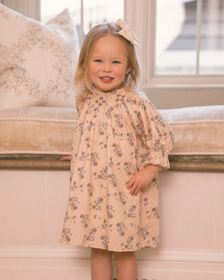 A young girl with blonde hair, wearing the noralee maddie dress in french floral—a vintage-inspired piece adorned with delicate floral embroidery and a large bow—smiles while standing indoors in front of a cushioned window seat.