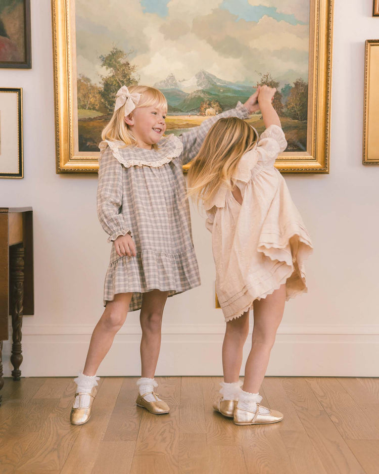 Two young girls in vintage-inspired outfits—one twirling in the noralee noelle dress in fall plaid, the other wearing a similar charming style with gold shoes—dance joyfully hand-in-hand before a wall lined with large, framed landscape paintings, their smiles glowing under warm light on the wooden floor.