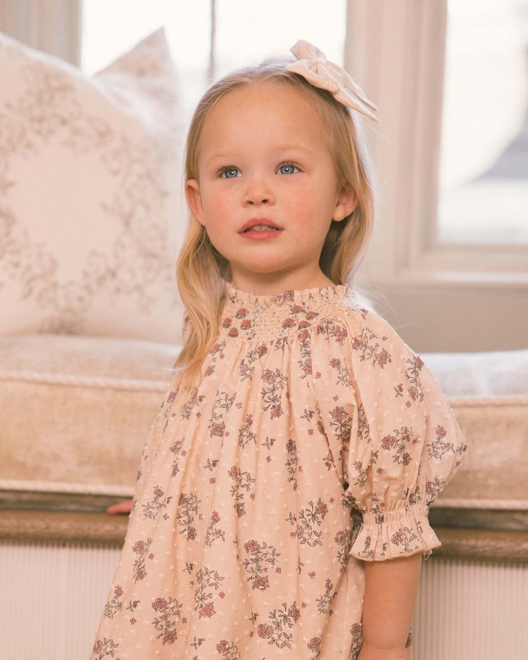A young girl with long blond hair stands indoors by a window, softly lit, with a cream cushion behind her. She wears the noralee “maddie dress in french floral,” a light pink, vintage-inspired piece crafted from delicate swiss dot fabric and accented with subtle floral embroidery. Looking slightly upward, she maintains a neutral expression.