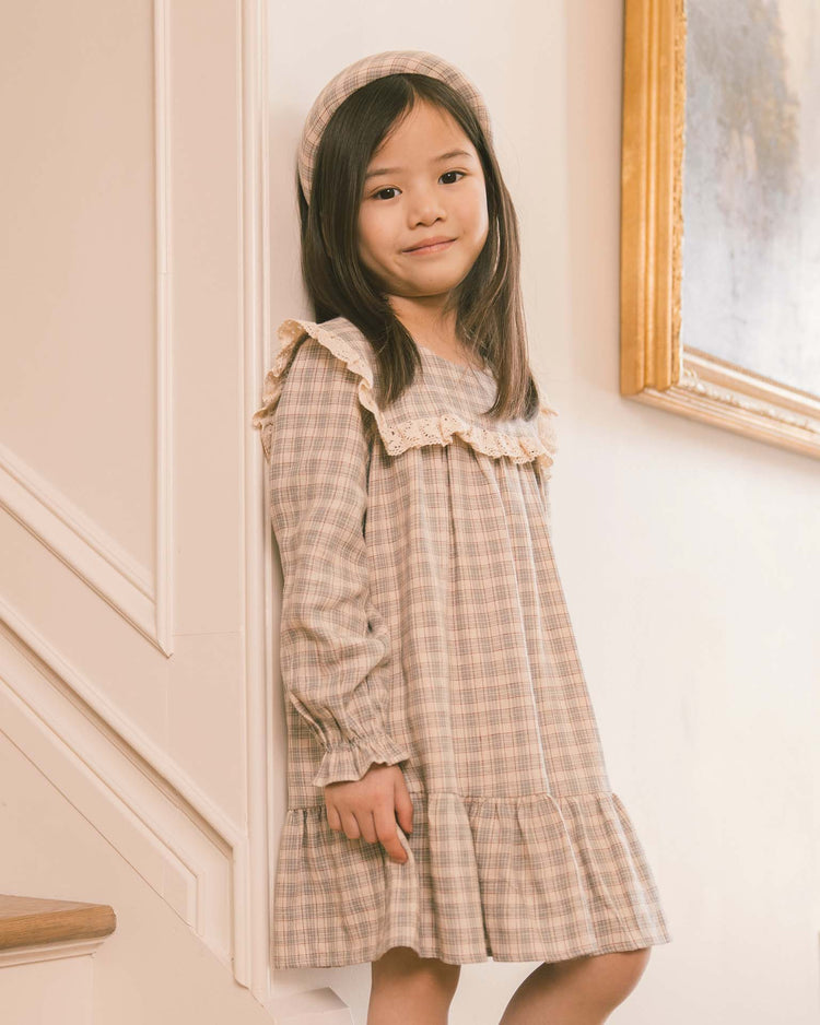 A young girl with long dark hair stands by a staircase indoors, smiling gently, dressed in the noralee noelle dress in fall plaid, which features a charming sailor bib collar. A framed painting hangs on the wall beside her.