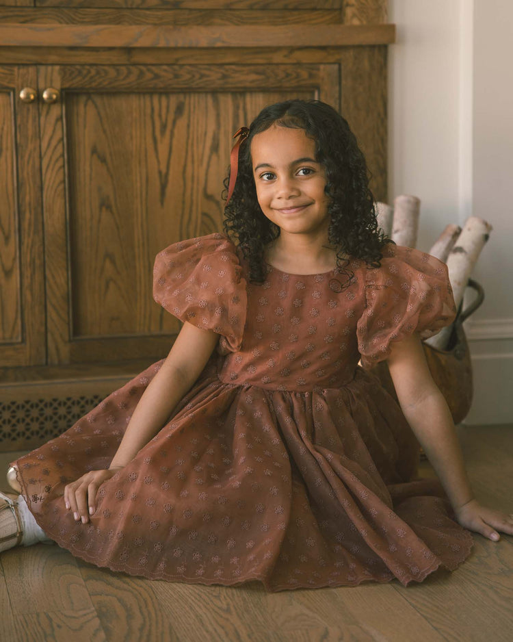 A young girl with curly hair, wearing the noralee sofia dress in spice floral—with balloon puff sleeves—sits smiling on a wooden floor. Behind her is a wooden cabinet and a basket with birch logs.