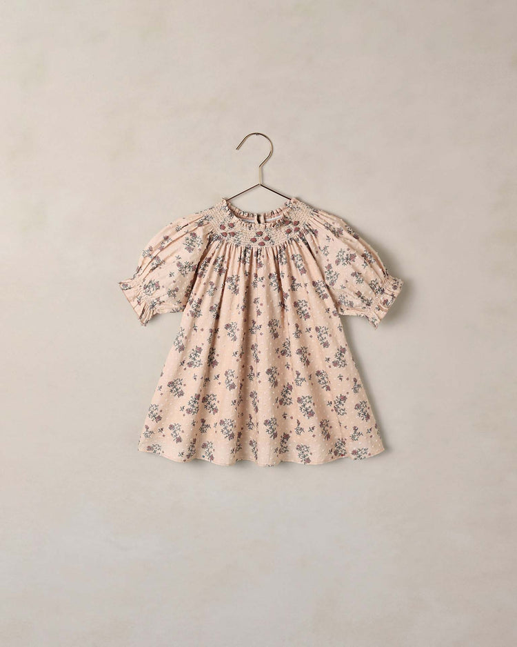 The maddie dress in french floral by noralee—a light pink baby dress adorned with a charming floral print, crafted from delicate swiss dot fabric with short puff sleeves and smocking at the neckline—hangs on a simple wire hanger against a neutral background.