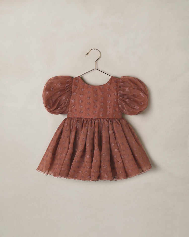 The sofia dress in spice floral by noralee—a brown, short-sleeved organza dress with balloon puff sleeves, a flared skirt, subtle floral patterns, and delicate flower eyelet details—hangs on a gold hanger against a light beige background.