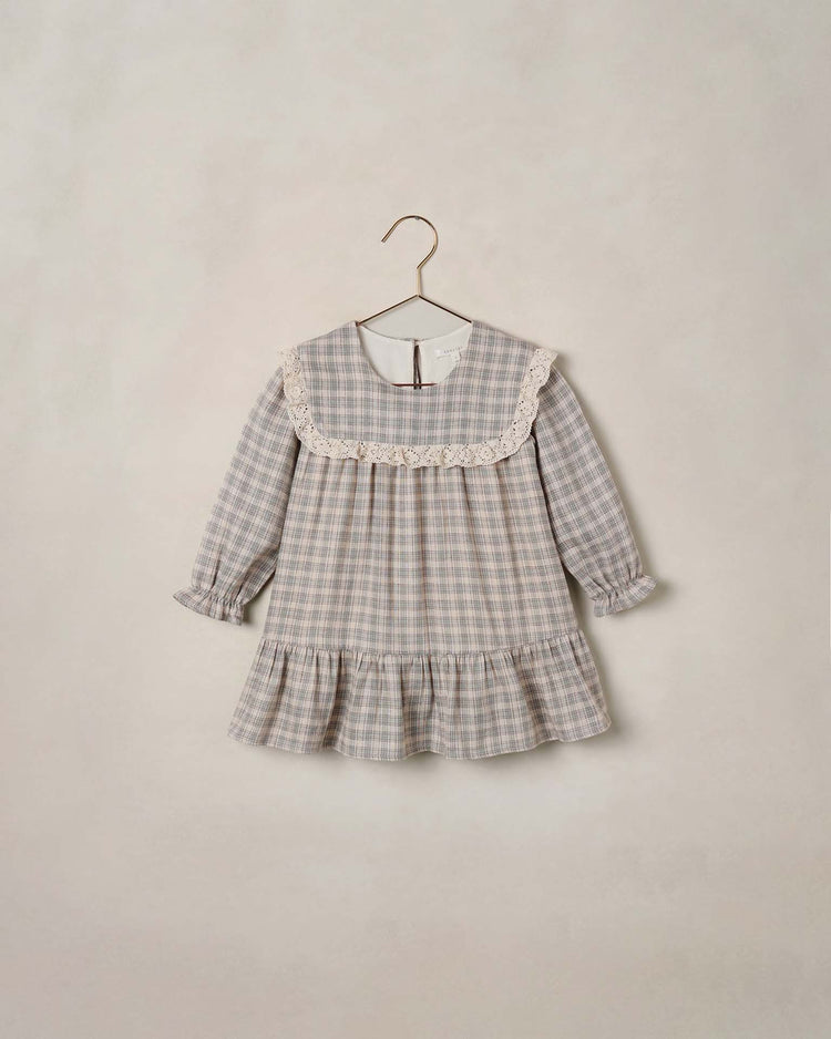 The noelle dress in fall plaid by noralee is a delightful autumn plaid dress for children, showcasing ruffled cuffs, a delicate lace sailor bib collar inspired by the signature Noelle Dress design, and a ruffled hem. It is elegantly displayed on a minimalist wire hanger against a light beige background.