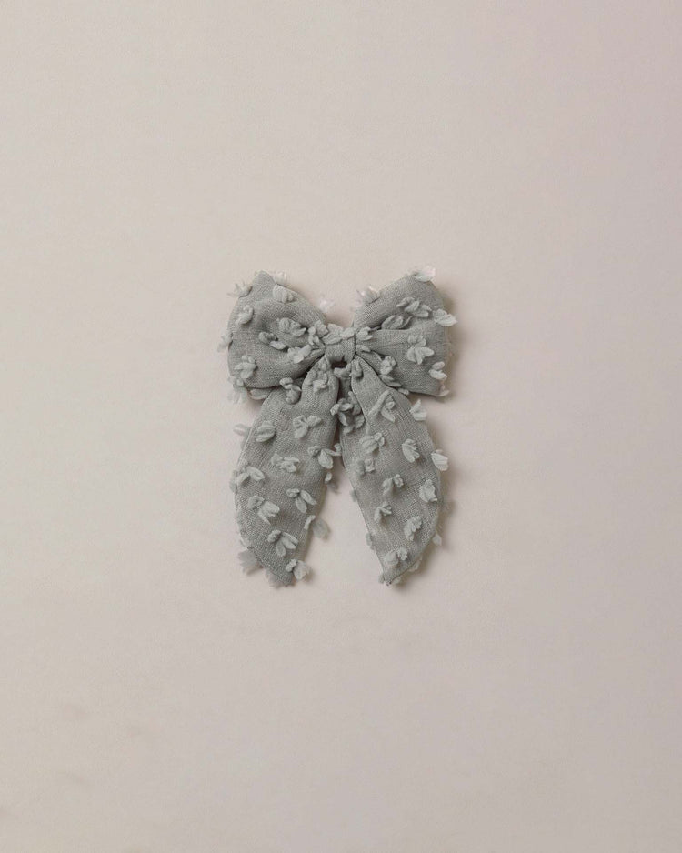 The everly bow in eucalyptus by noralee, featuring a soft eucalyptus hue with small textured dots and frayed edges, is displayed against a simple light background; its gray tone and secure alligator clip lend the design a delicate charm.
