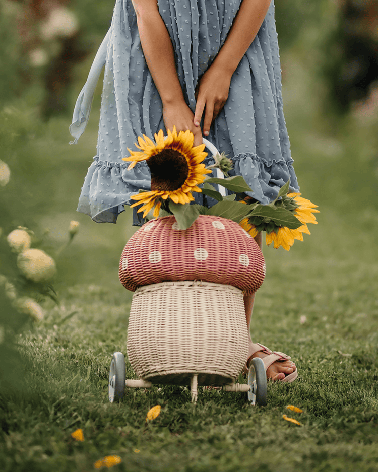 A child in a blue dress stands on grass, holding sunflowers and the dinkum dolls rattan mushroom luggy in musk—an adorable wheeled basket for toy storage—outdoors among green plants and flowers.