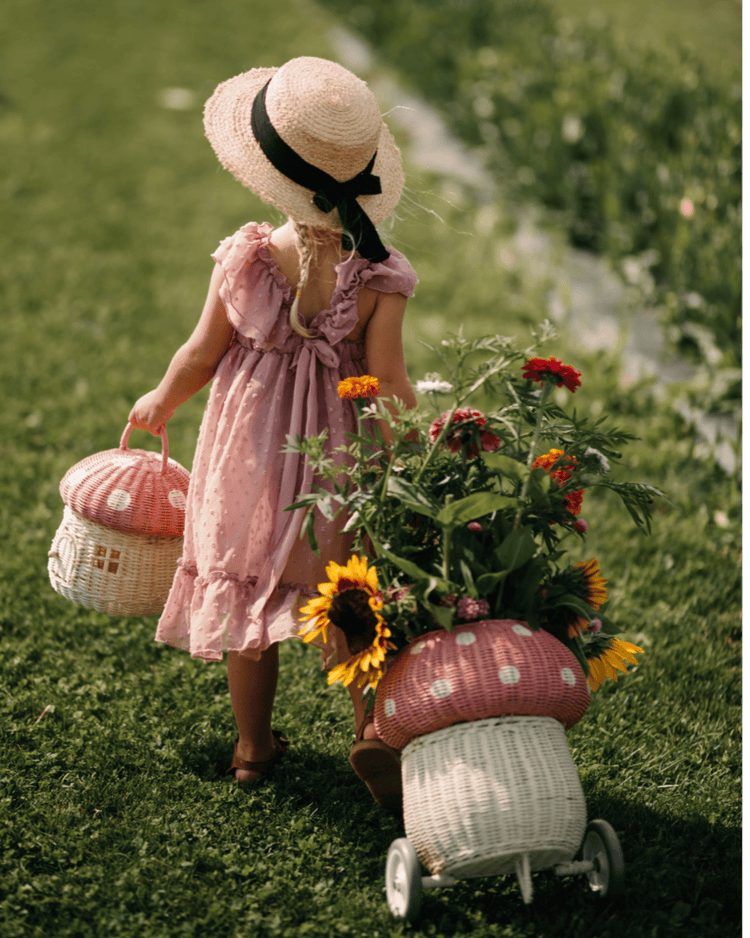 A girl in a pink dress and straw hat walks on grass, carrying a basket and pulling the dinkum dolls rattan mushroom luggy in musk filled with sunflowers on a sunny day.