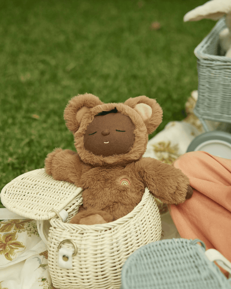 A dinkum dolls plush toy with brown fur and bear ears rests inside a mini chari bag in chalk on a picnic blanket, with grass in the background.