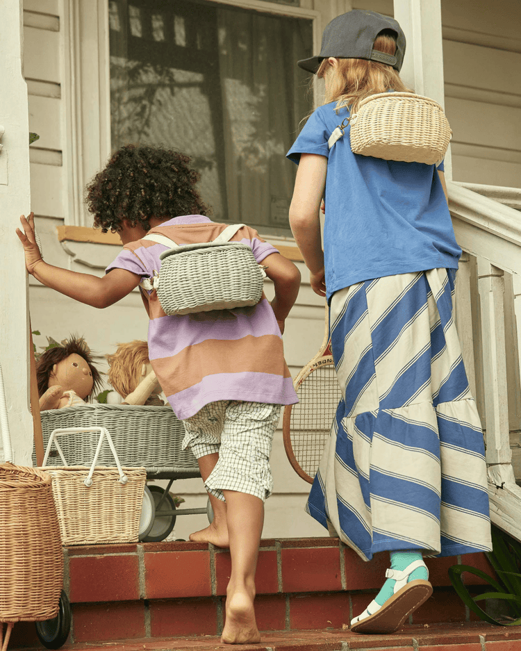 Two children climb the porch steps, each carrying a dinkum dolls mini chari bag in vintage blue on their backs. One wears a blue shirt and striped skirt; the other, a striped shirt and plaid shorts. A cart with dolls sits nearby.