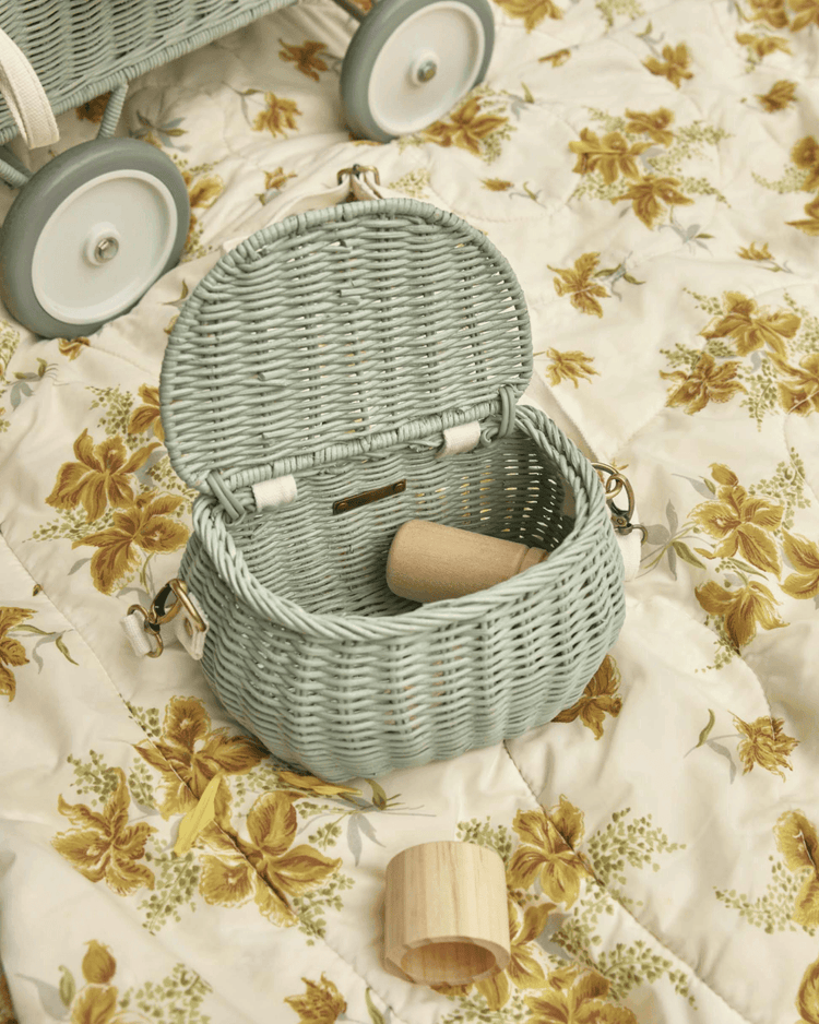 A dinkum dolls mini chari bag in vintage blue sits open on a floral quilt, holding wooden toys. A gray wagon with white wheels is partly visible in the background.