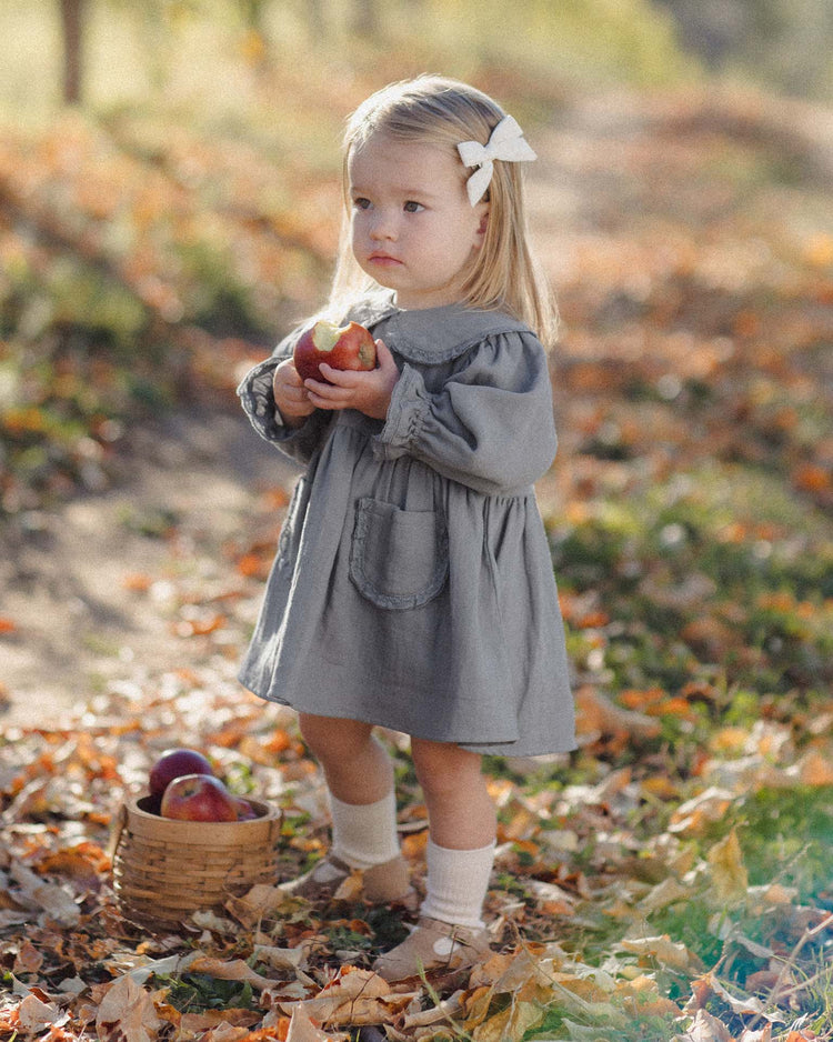 A young girl stands outdoors on a leaf-strewn path, holding apples, dressed in the marisol dress in slate by quincy mae. The dress features a Peter Pan collar and is made from soft organic cotton—ideal for an autumn day. She wears a white bow in her hair, and beside her on the ground rests a small basket of apples.