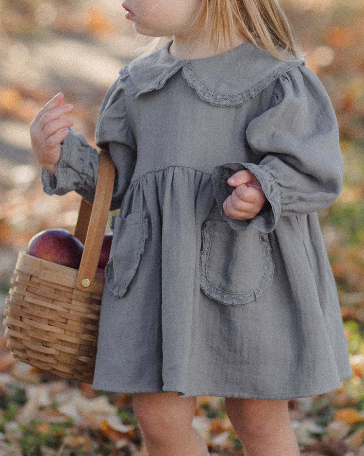 A young child wears the quincy mae marisol dress in slate, featuring a Peter Pan collar, while holding a small basket of apples outdoors among autumn leaves. The child's face is not visible.