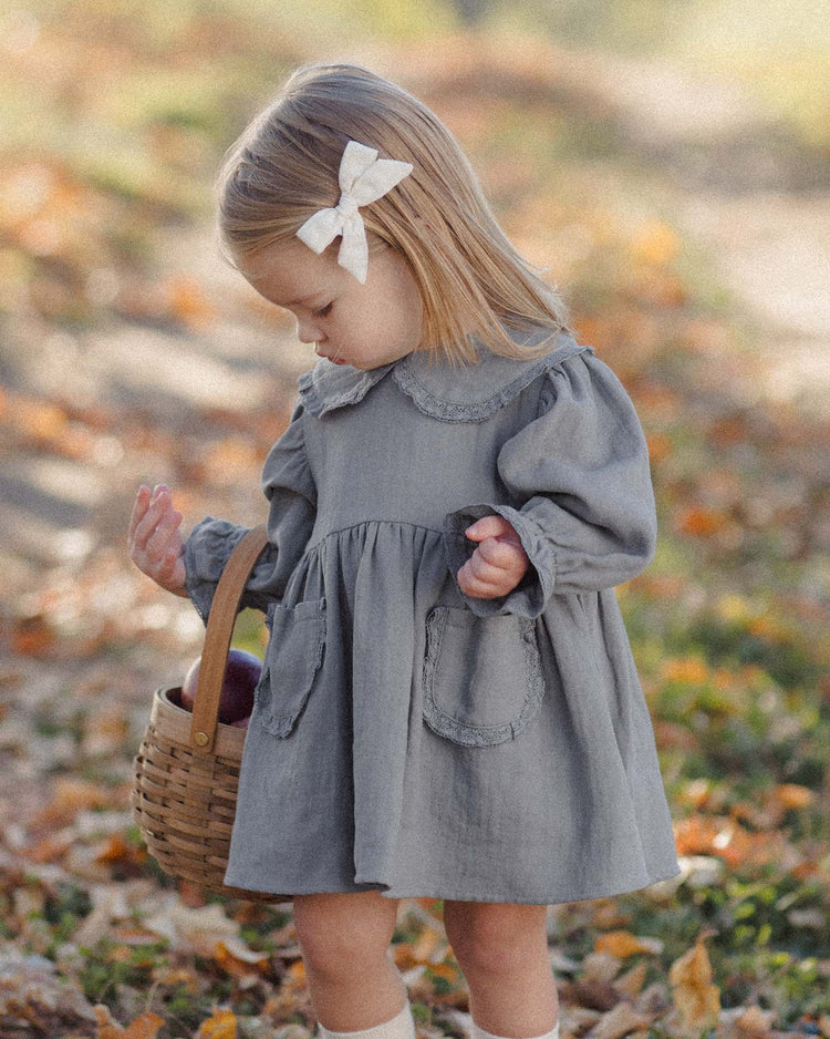 A young child wears the quincy mae marisol dress in slate, featuring a charming Peter Pan collar and white bow, while standing outdoors with a woven basket. Fallen leaves cover the ground, and the softly blurred background is filled with autumn colors.