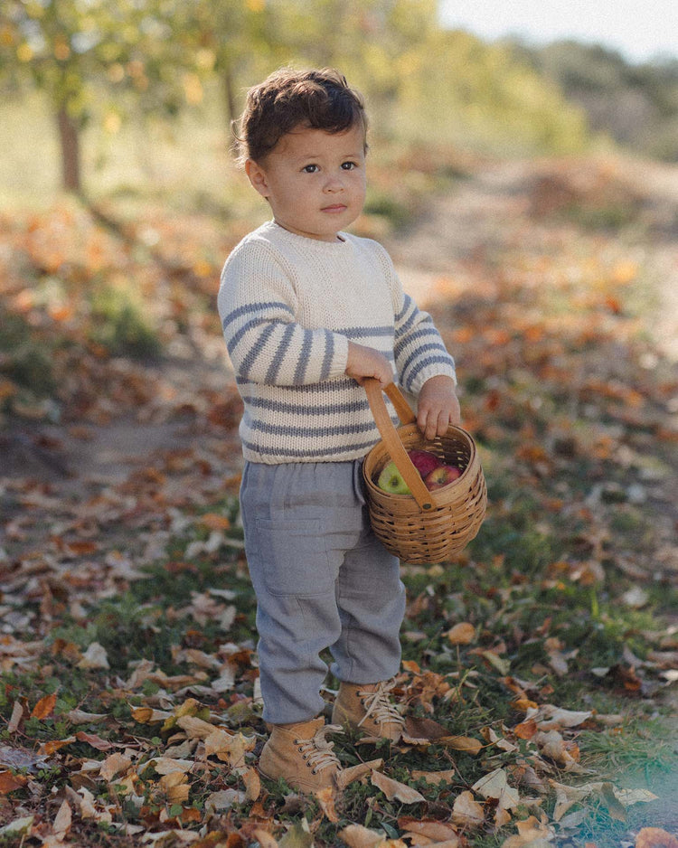 A young child stands outdoors on a leafy path, holding a small basket of apples. The child wears the quincy mae rex sweater in slate stripe, paired with gray pants and brown boots. Trees with green and yellow leaves are in the background.
