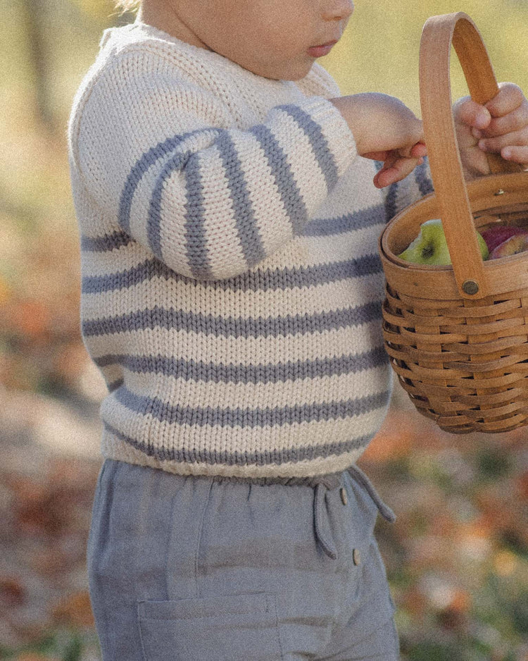 A young child wearing the quincy mae rex sweater in slate stripe and gray pants holds a wooden basket filled with apples outdoors in soft sunlight.