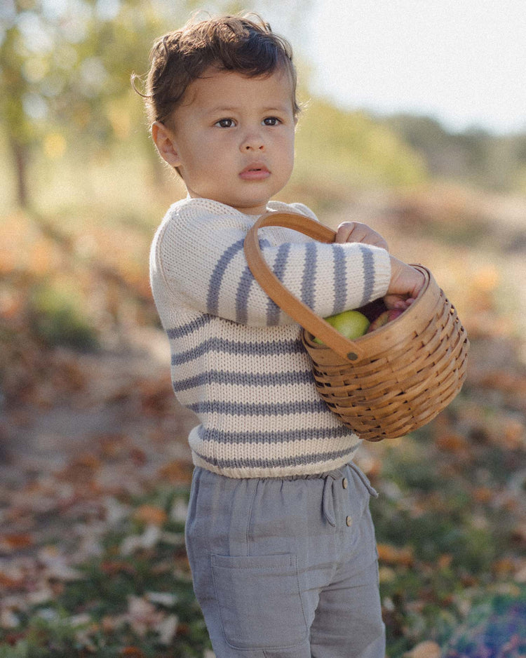 A young child wearing the quincy mae rex sweater in slate stripe holds a woven basket of apples outdoors, surrounded by autumn leaves and soft sunlight.